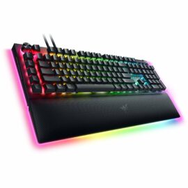 Das Bild zeigt eine schräge Draufsicht auf die Razer BlackWidow V4 Pro Gaming-Tastatur, die auf einer flachen Oberfläche liegt. Die Tastatur ist hintergrundbeleuchtet mit einer RGB-Beleuchtung, die in verschiedenen Farben leuchtet. An der Oberseite links befindet sich ein multifunktionaler Drehregler und zusätzliche Medientasten sind sichtbar. Das Bild hebt das gesamte Design und die visuellen Merkmale der Tastatur hervor, einschließlich der Handballenauflage mit einer strukturierten Oberfläche am unteren Rand, und dient dazu, die Tastatur für Marketing- oder Verkaufszwecke ansprechend zu präsentieren.